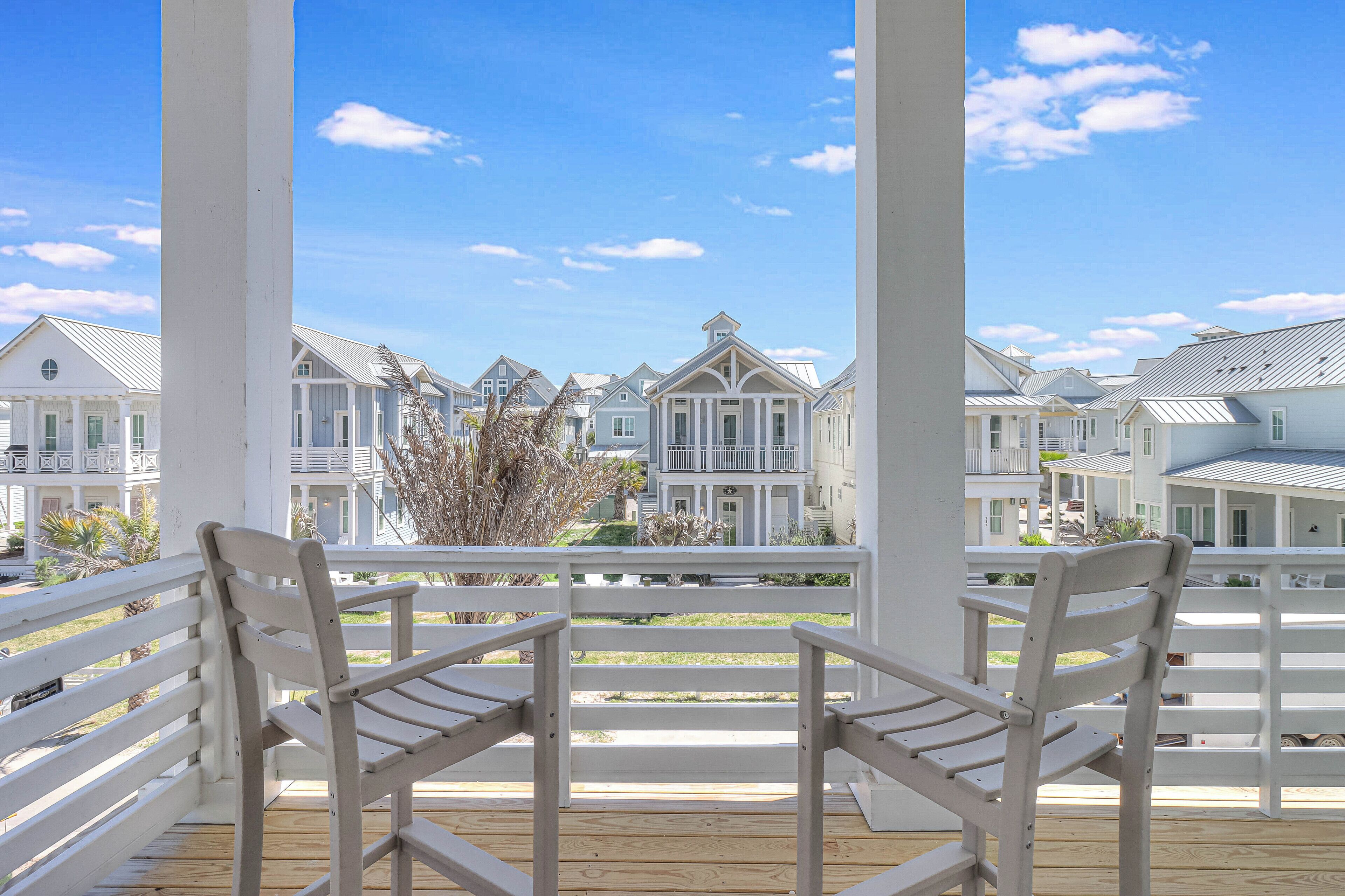 A wooden deck with white railings and two white chairs facing a row of white houses with gabled roofs and multiple windows.
