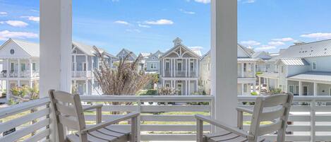 A wooden deck with white railings and two white chairs facing a row of white houses with gabled roofs and multiple windows.