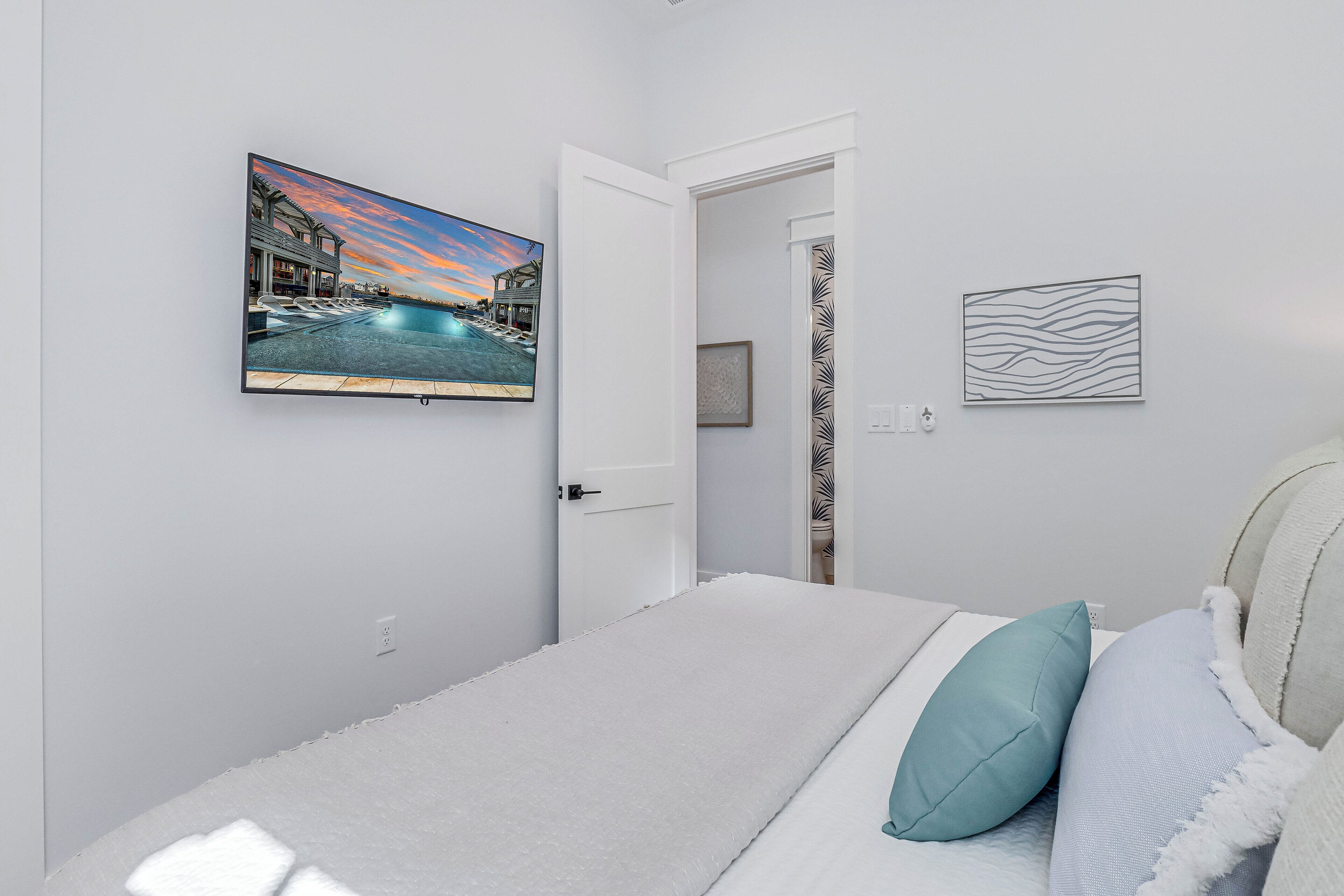 Un dormitorio con cama, almohadas, un televisor de pared y una puerta que da a otra habitación.