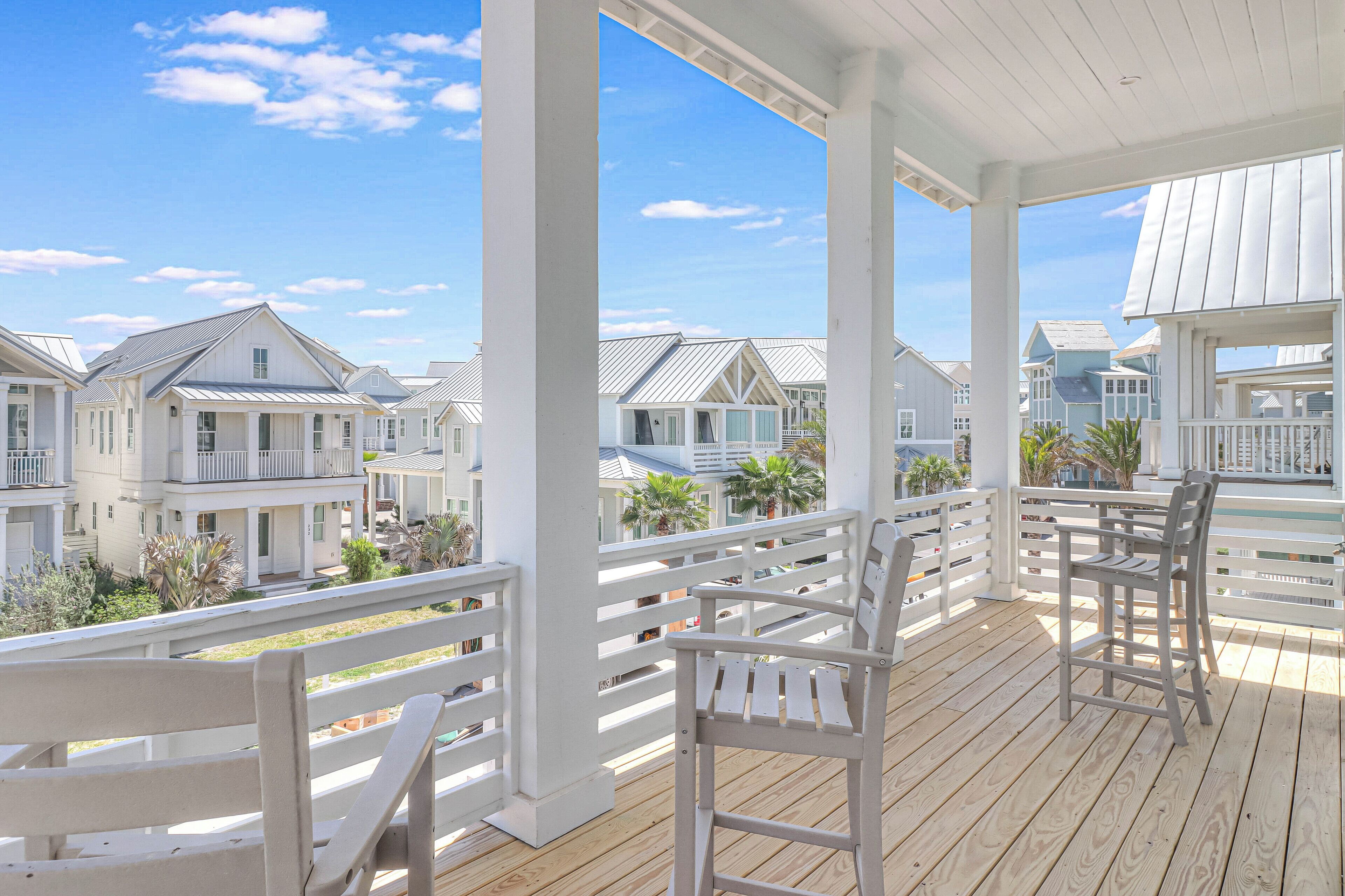 A wooden deck with white railings and chairs, overlooking a row of beach houses with white exteriors and blue roofs.