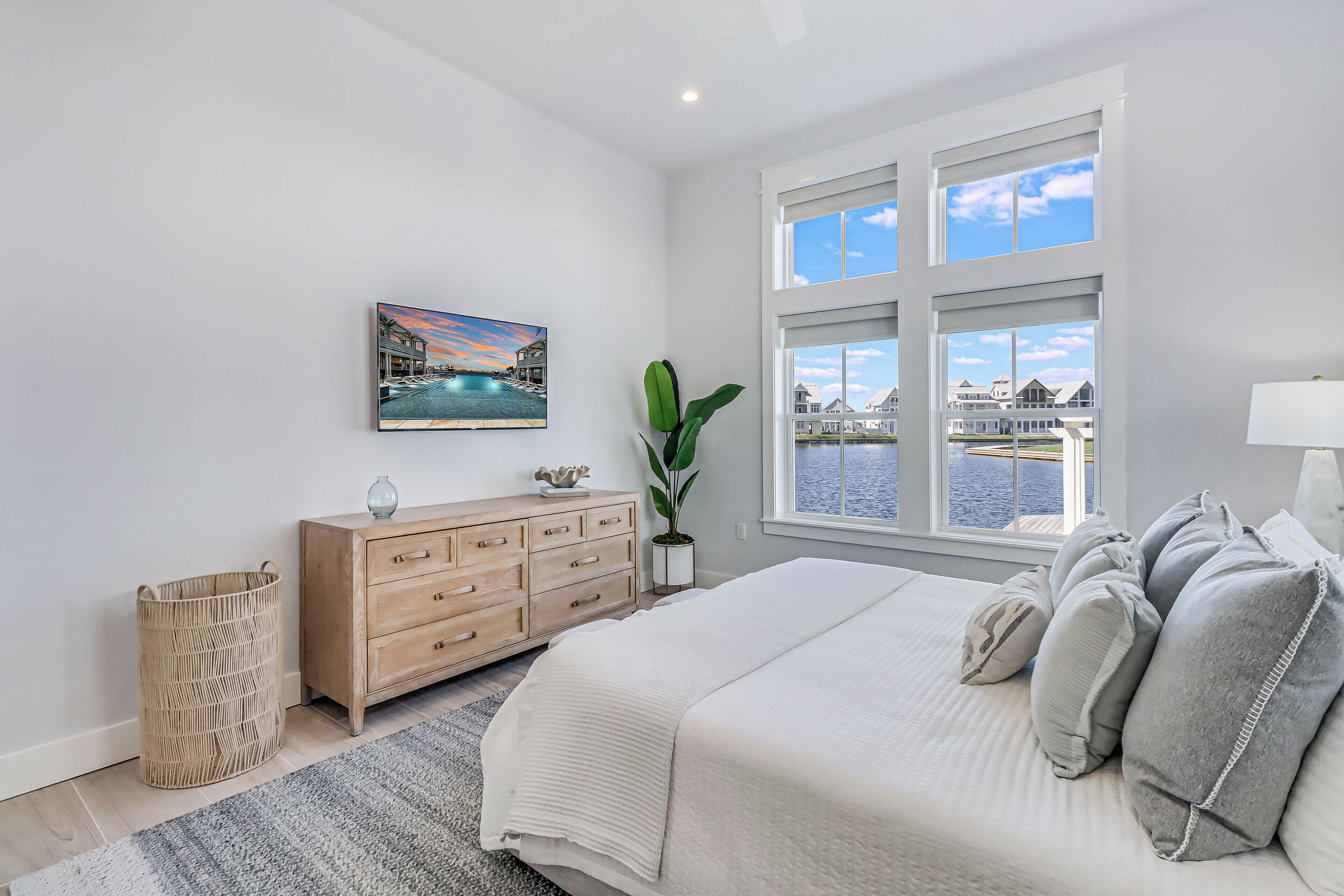 A bedroom with a large bed, a wooden dresser, a plant, a painting, and a view of the water through the window.