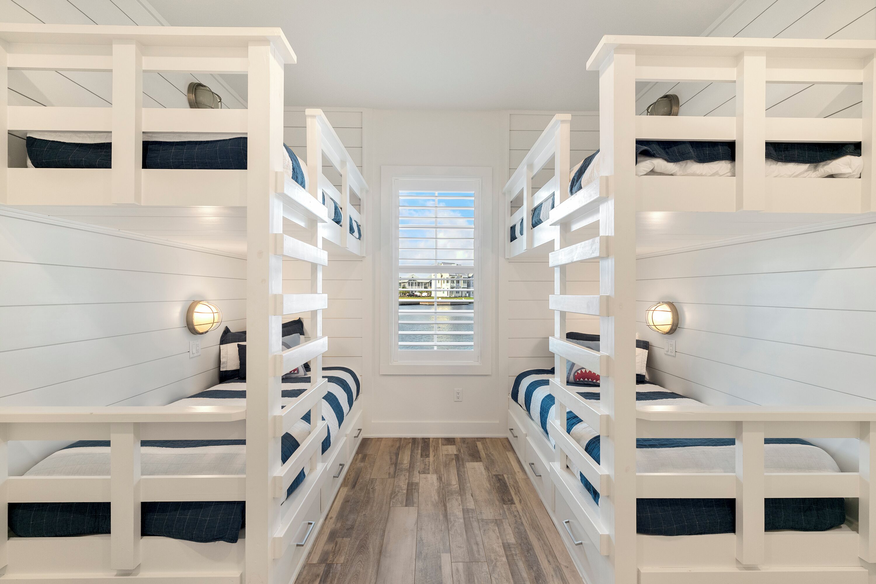 A narrow hallway with bunk beds on either side, a central window, and wall-mounted light fixtures.