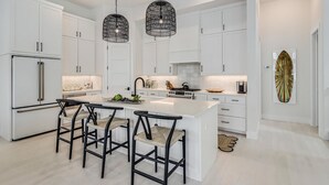 A modern kitchen with white cabinetry, a central island with bar stools, pendant lighting, and a surfboard decoration on the wall. - Friday's Comin' 132 BAR (Port Aransas)