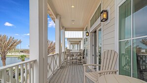 A covered outdoor area with a wooden deck, chairs, and tables, overlooking a body of water and palm trees. - Friday's Comin' 132 BAR (Port Aransas)