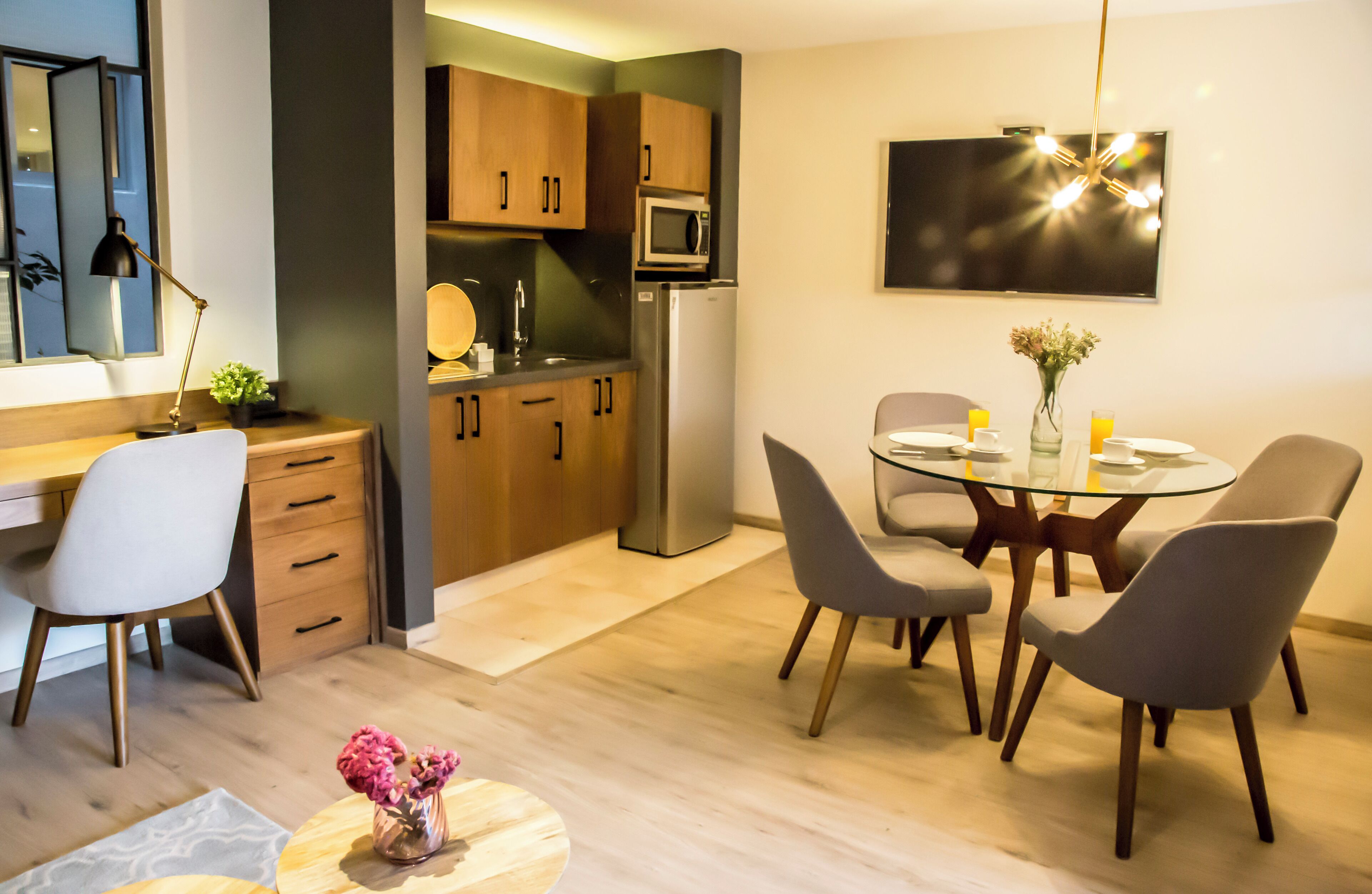 Une cuisine moderne avec des meubles en bois, un four à micro-ondes et un réfrigérateur. On y trouve un coin repas avec une table et des chaises en verre, ainsi qu'une petite plante posée sur la table.