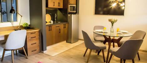 Une cuisine moderne avec des meubles en bois, un four à micro-ondes et un réfrigérateur. On y trouve un coin repas avec une table et des chaises en verre, ainsi qu'une petite plante posée sur la table.
