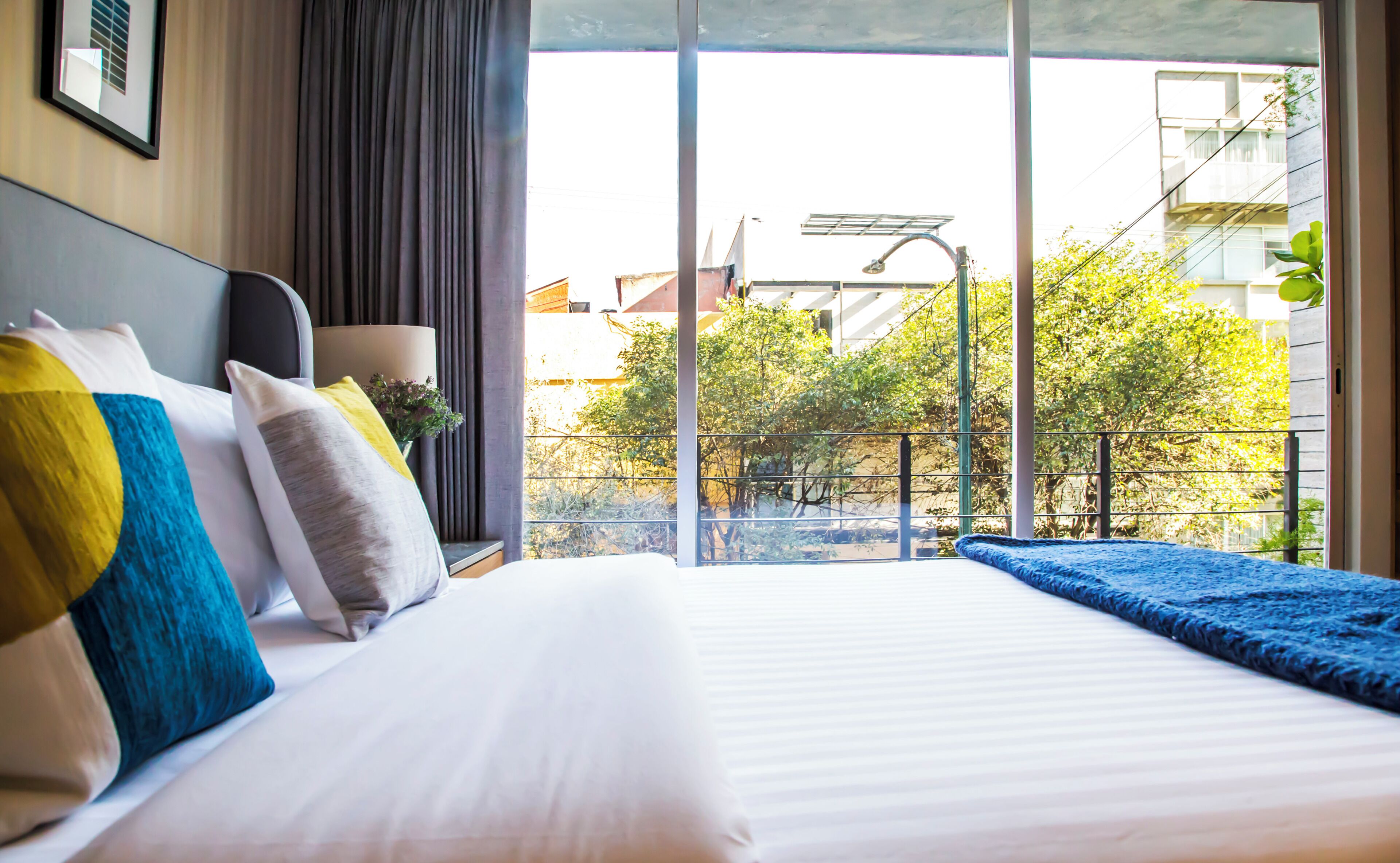 A hotel room with a large bed, a window with a view of buildings and trees, and a balcony with a railing.