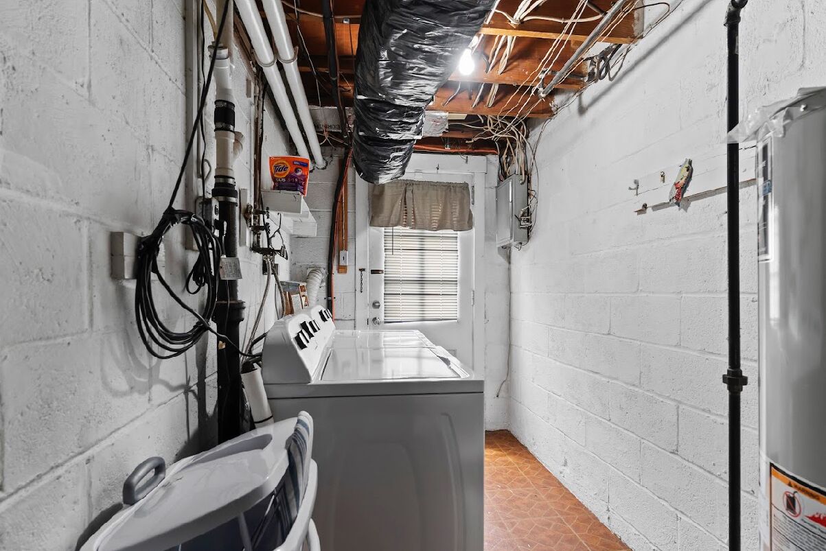 A laundry room with a washing machine, a dryer, and a utility sink. There are exposed pipes and wires on the ceiling, and a window with blinds.