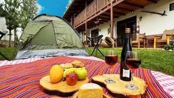 Una carpa montada sobre una manta de rayas coloridas, con una mesa de comida y bebidas, una terraza de madera y un edificio al fondo.