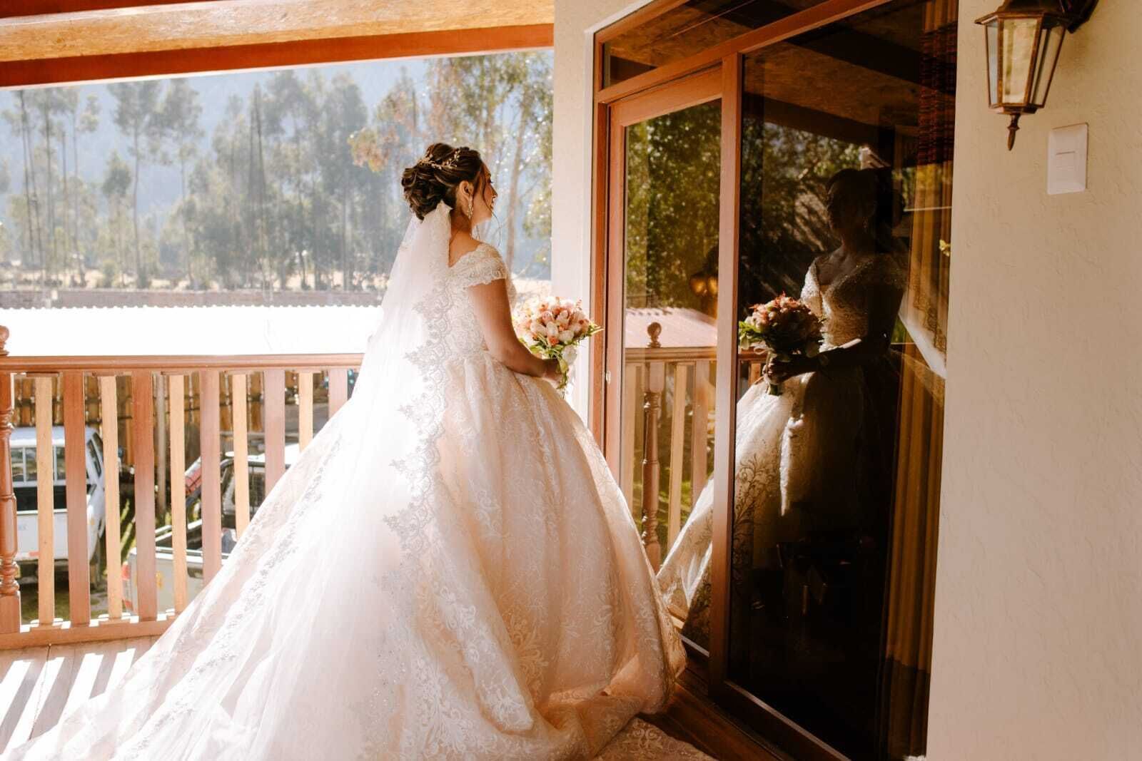 Una novia con vestido de novia blanco de pie en un balcón con vista a árboles y coches aparcados.