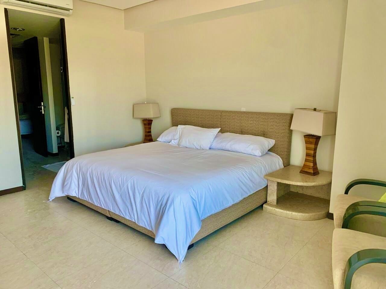 A bedroom with a bed, two bedside tables with lamps, a chair, and a door leading to another room.