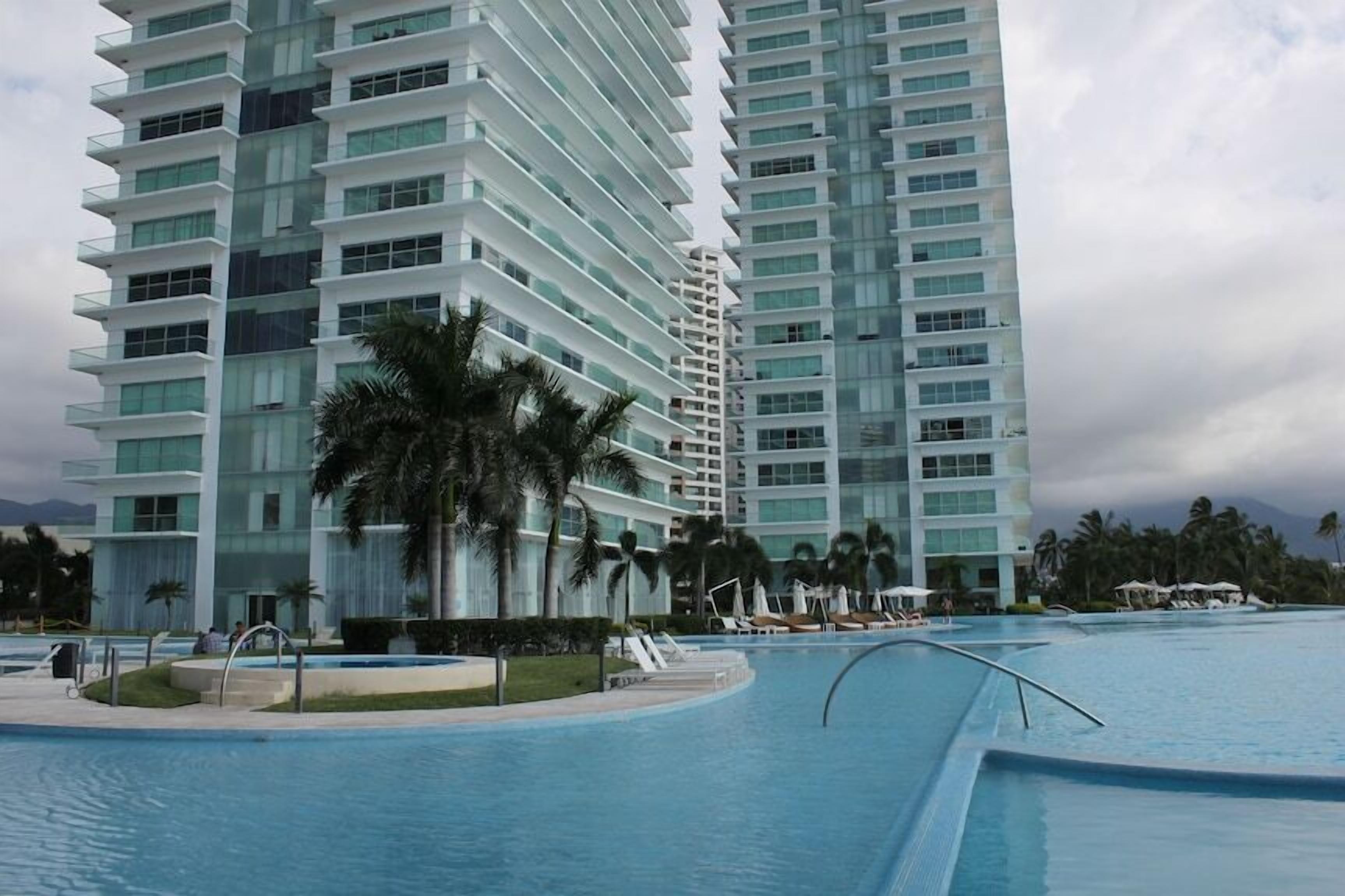 A modern hotel with a large swimming pool, palm trees, and high-rise buildings in the background.