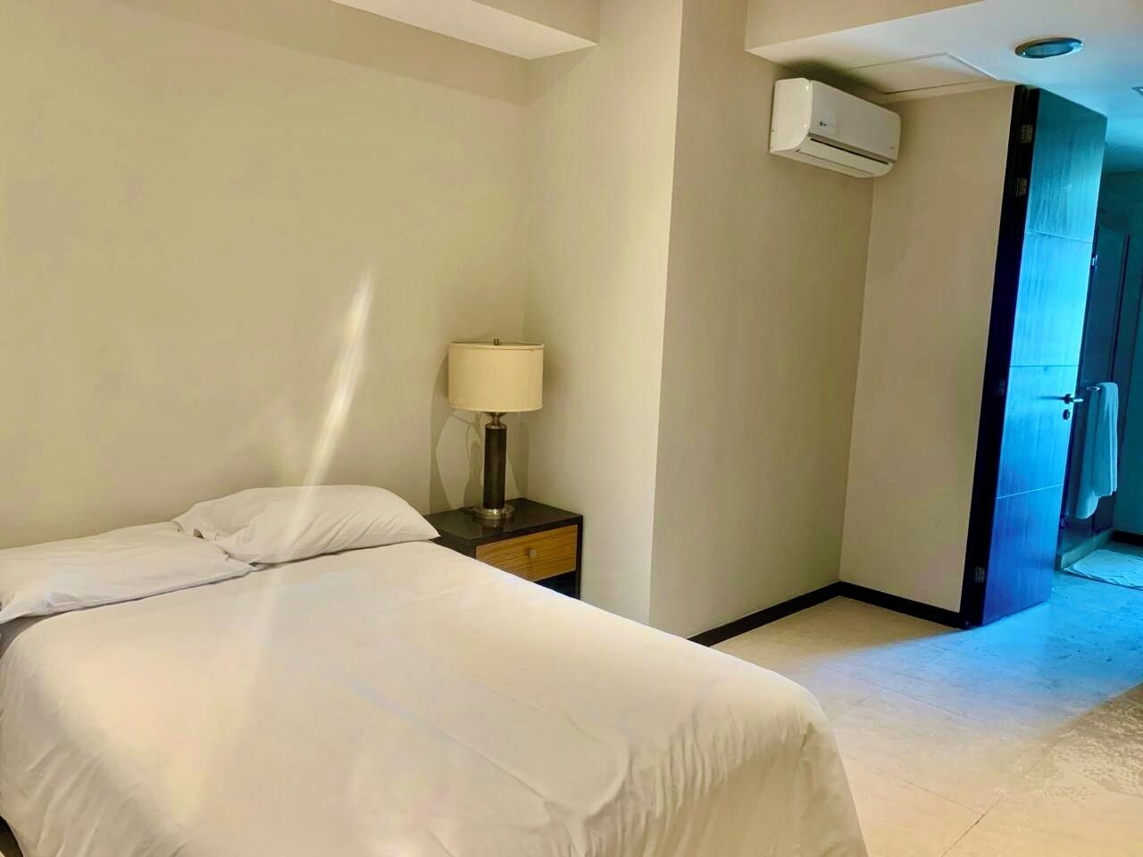 A hotel room with a bed, a nightstand with a lamp, an air conditioning unit, and a door leading to another room.