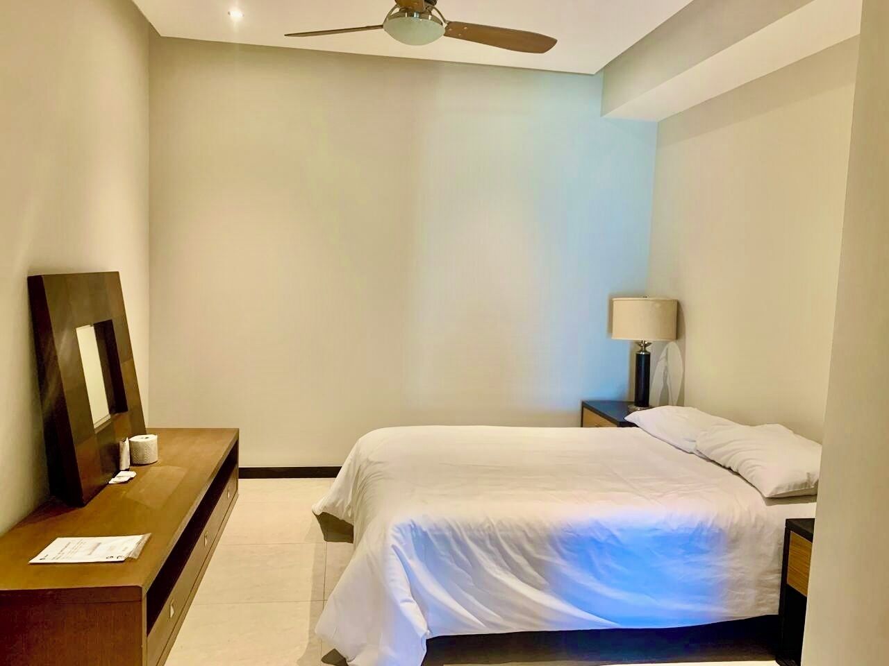 Habitación de hotel con cama, mesita de noche con lámpara, cómoda de madera y ventilador de techo.