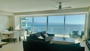 Una sala de estar moderna con un sofá negro grande, una zona de comedor con sillas blancas y vista al océano a través de ventanas amplias.