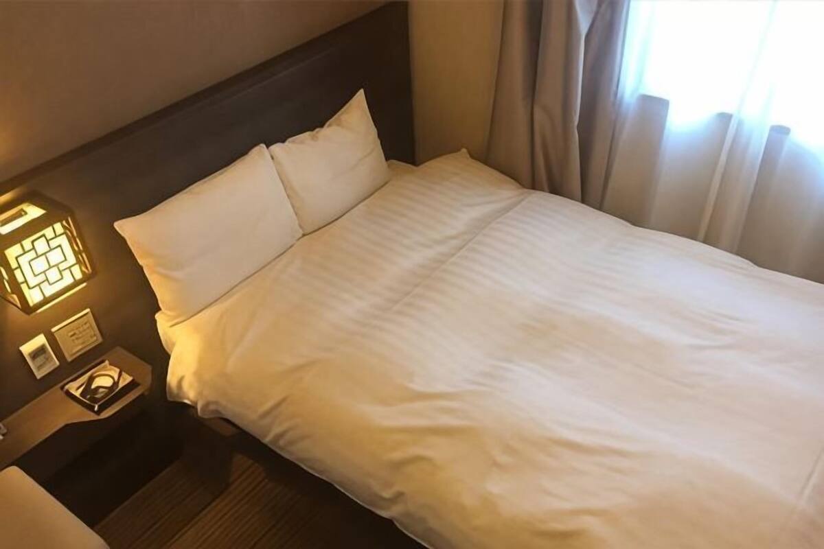 A hotel room with a neatly made bed, a nightstand with a lamp, and a window with curtains.