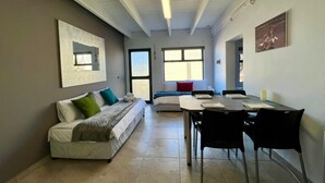 Una habitación moderna con una cama, un escritorio y sillas. Cuenta con una ventana que proporciona luz natural y una puerta que da a otra habitación.