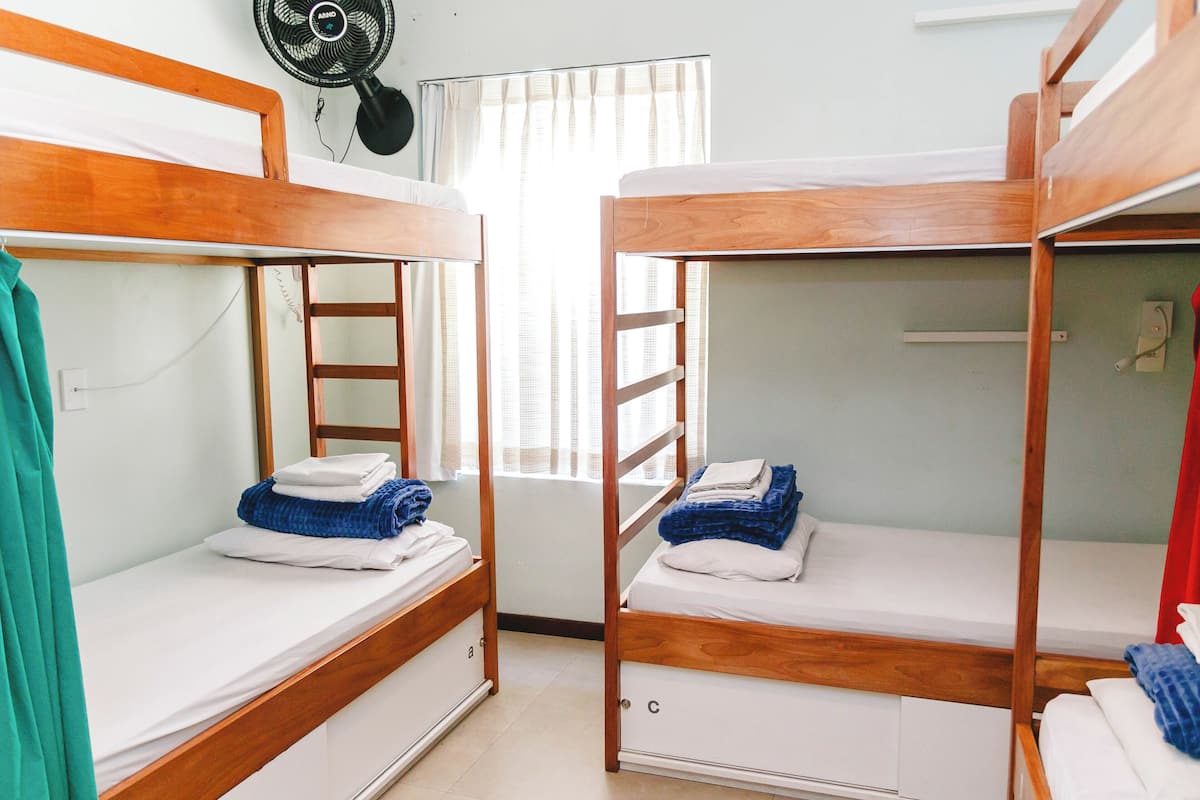 A dormitory room with bunk beds, a fan, and a window with curtains.