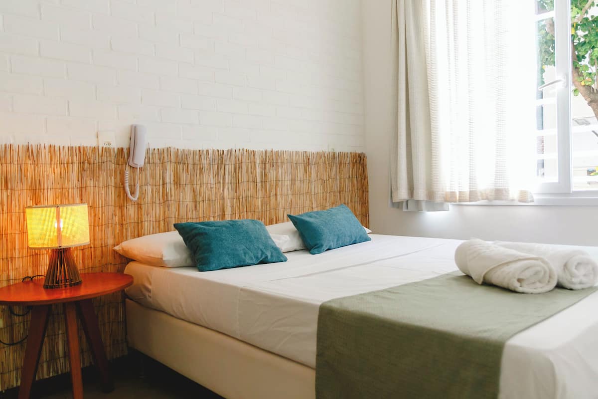 A hotel room with a white brick wall, a bamboo headboard, a bed with white linens and teal pillows, a bedside lamp, and a window with curtains.