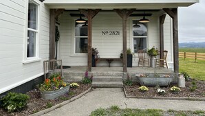 A white house with the number 2821 on the front porch, featuring a covered seating area with two chairs and a table, surrounded by flower beds and a small garden. - Farmhouse in beautiful Ferndale. Big enough for the whole family! (Ferndale)