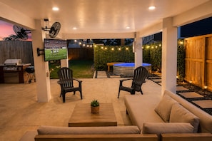 An outdoor patio area with a TV screen, two Adirondack chairs, a sofa, a hot tub, and a grill.