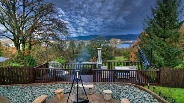A backyard with a barbecue grill, stone benches, a wooden deck, and a view of a lake and hills. - Gleddoch Coach House offers an exclusive retreat near Glasgow. (Langbank)