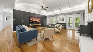 A spacious living room with a ceiling fan, a fireplace, and a large window offering a view of greenery.