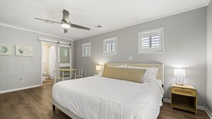 A bedroom with a large bed, a ceiling fan, a nightstand with a lamp, and a door leading to a bathroom.
