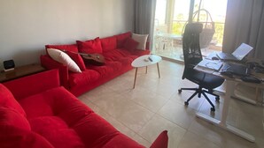 A modern living room with a red sectional sofa, a small white coffee table, and a black office chair. There is a balcony with a view of the outdoors. - Regina Gardens 1, Venus Beach -6 min, Sunset Sea View, Gated Complex (Paphos)