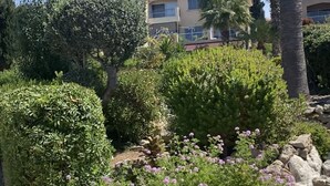 A landscaped garden with various plants and flowers, a stone pathway, and a building in the background. - Regina Gardens 1, Venus Beach -6 min, Sunset Sea View, Gated Complex (Paphos)
