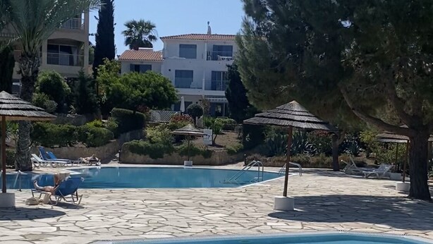 A pool area with stone flooring, lounge chairs, and umbrellas. There are buildings in the background. - Regina Gardens 1, Venus Beach -6 min, Sunset Sea View, Gated Complex (Paphos)