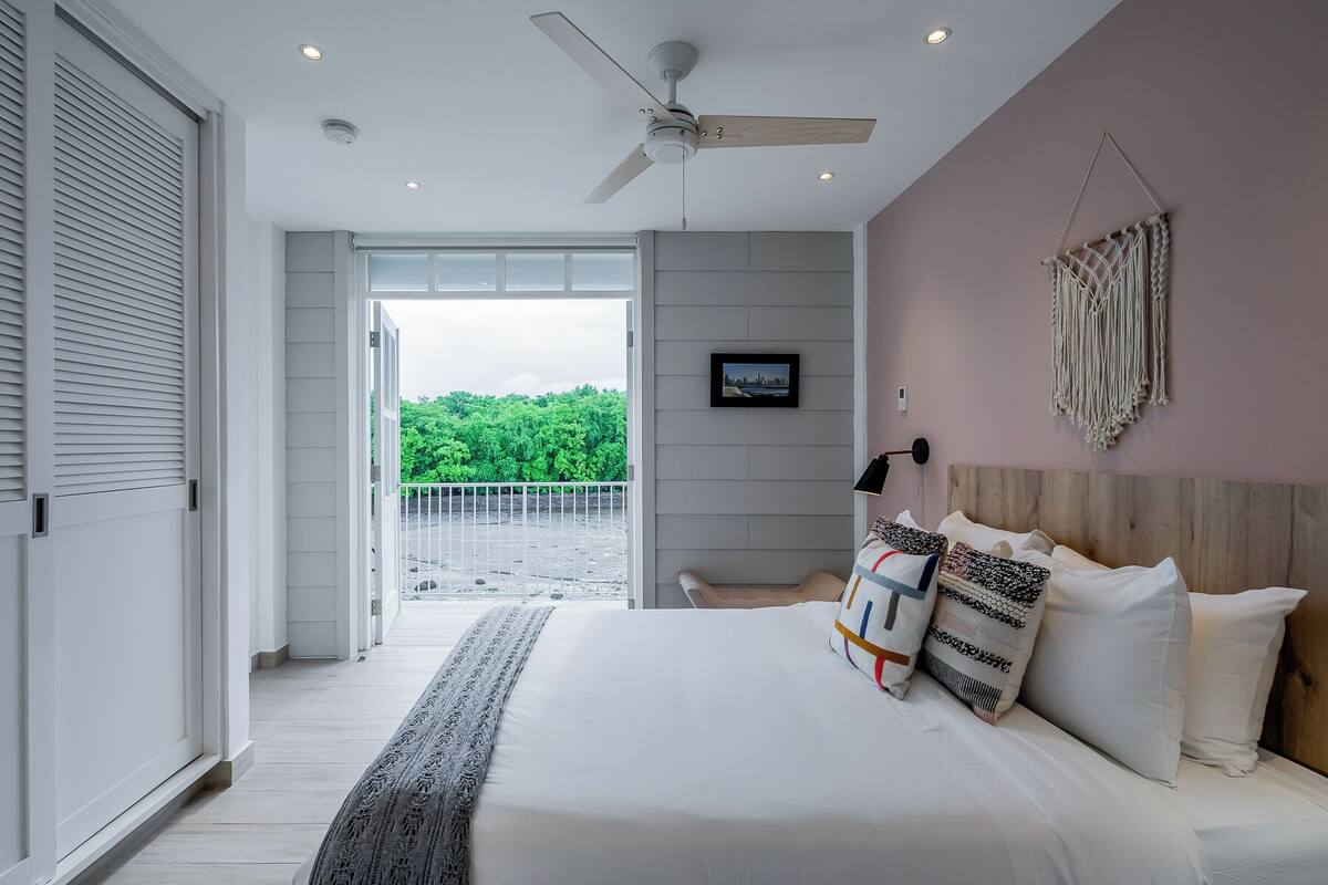 A bedroom with a large bed, a ceiling fan, a TV on the wall, and a view of greenery outside.