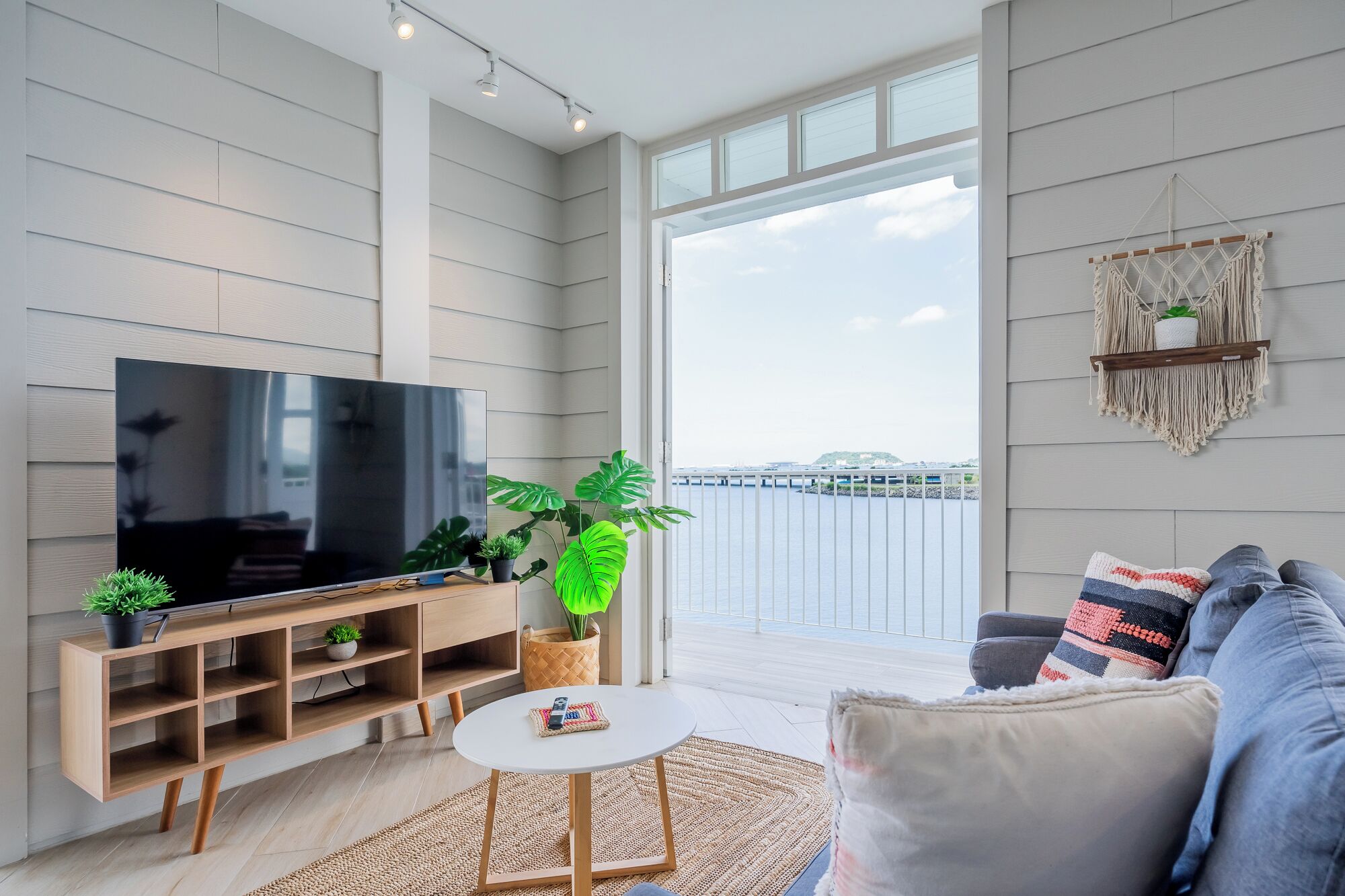 Ein modernes Wohnzimmer mit Fernseher, einer Holz-Unterhaltungsecke, Topfpflanzen, einem runden Couchtisch und einem grauen Sofa mit gemusterten Kissen.
