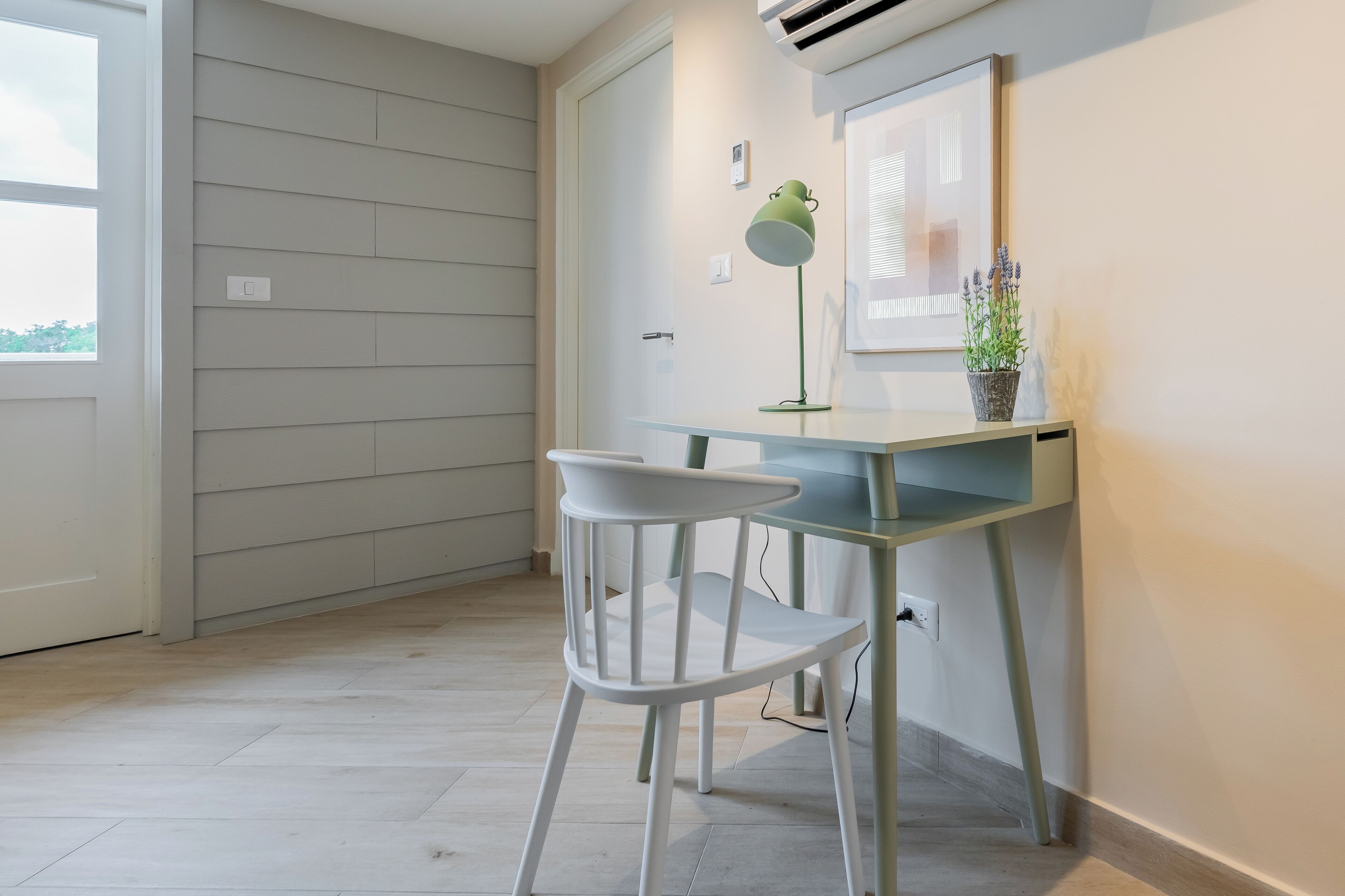 Une petite pièce moderne, équipée d’un bureau et d’une chaise, d’une lampe de bureau verte et d’une plante en pot.
