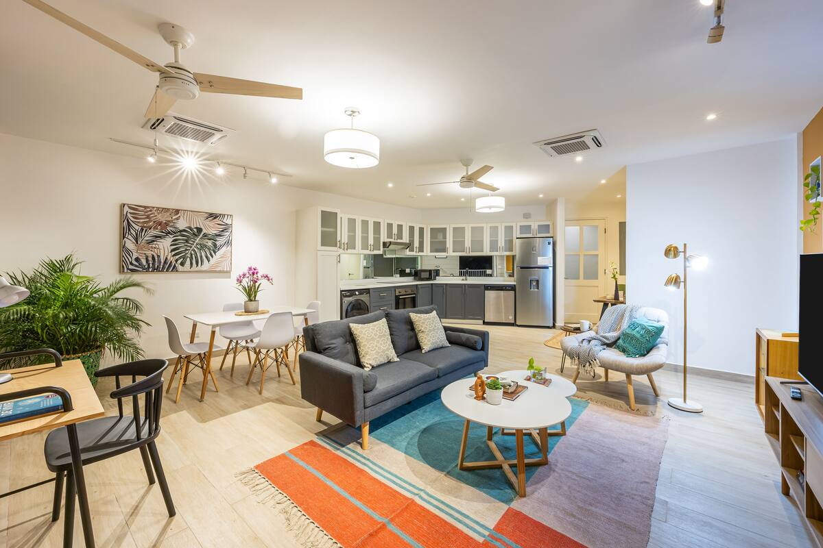 A modern living room with a gray sofa, dining table, and chairs, a kitchen in the background, and a ceiling fan.