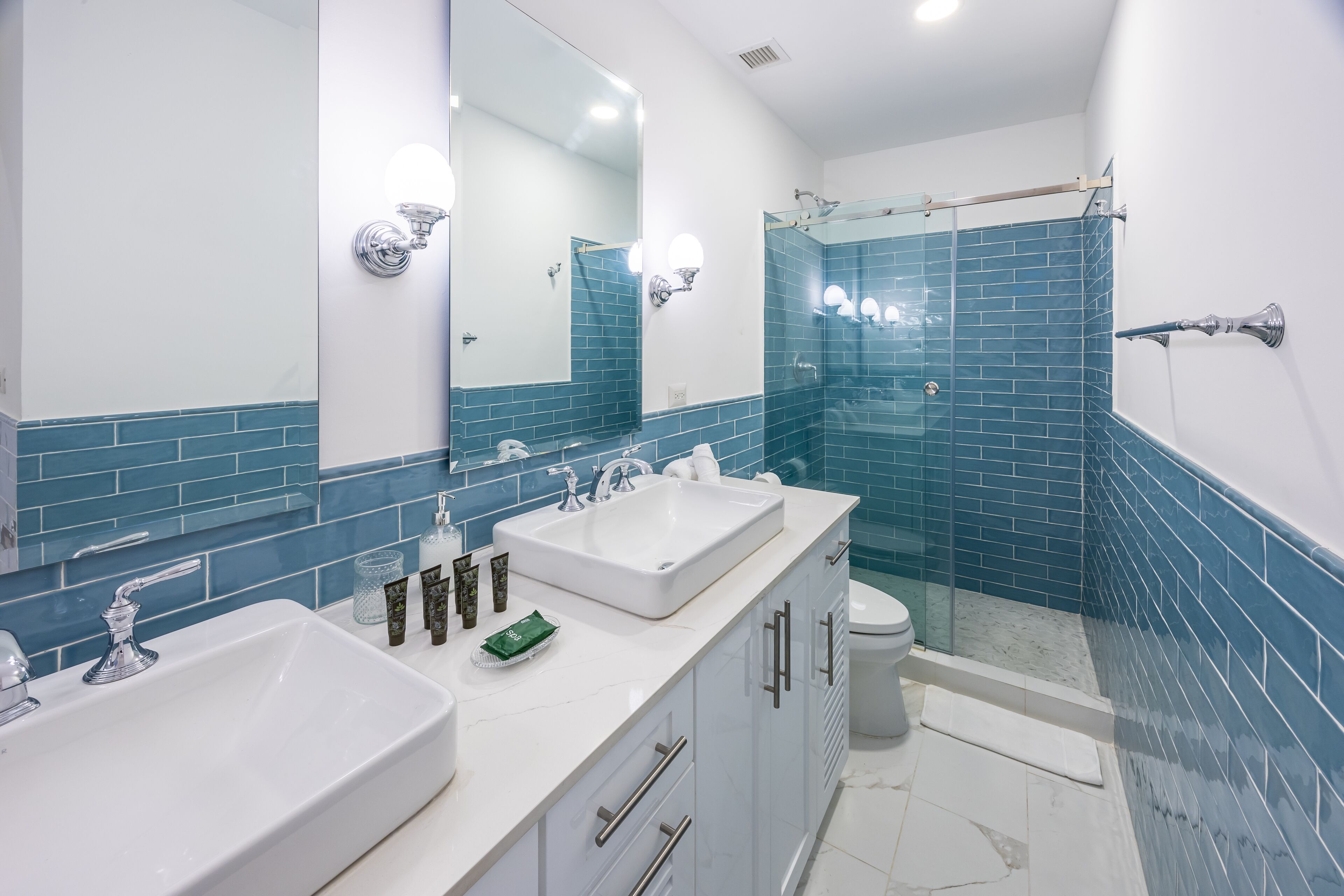 Un baño moderno con un espejo grande, dos lavamanos, una ducha y paredes con azulejos azules.