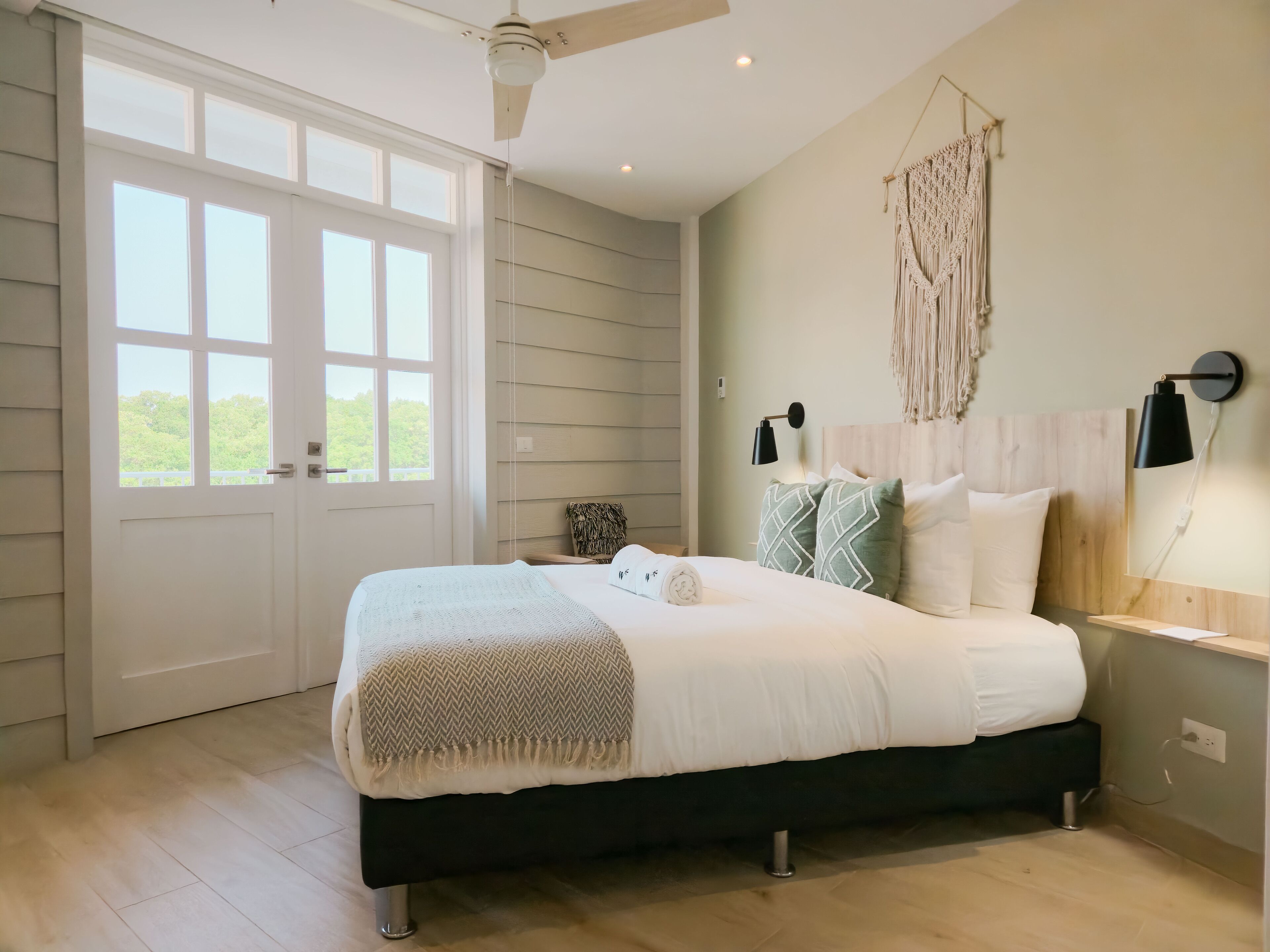 A bedroom with a large bed, a ceiling fan, a macrame wall hanging, and a view through a large window.