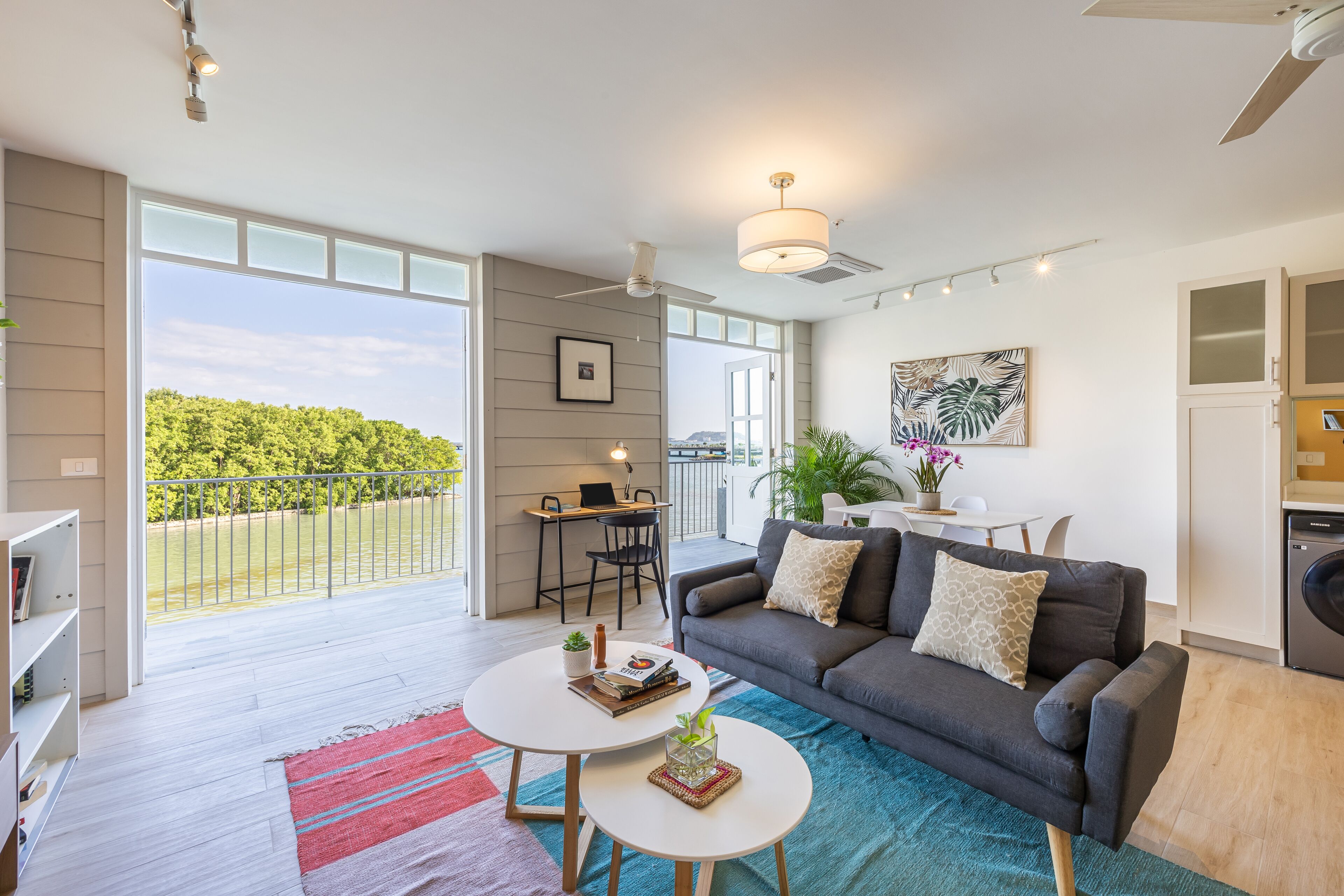 A modern living room with a grey sofa, a coffee table, and a dining area with a table and chairs. There is a balcony with a view of greenery.