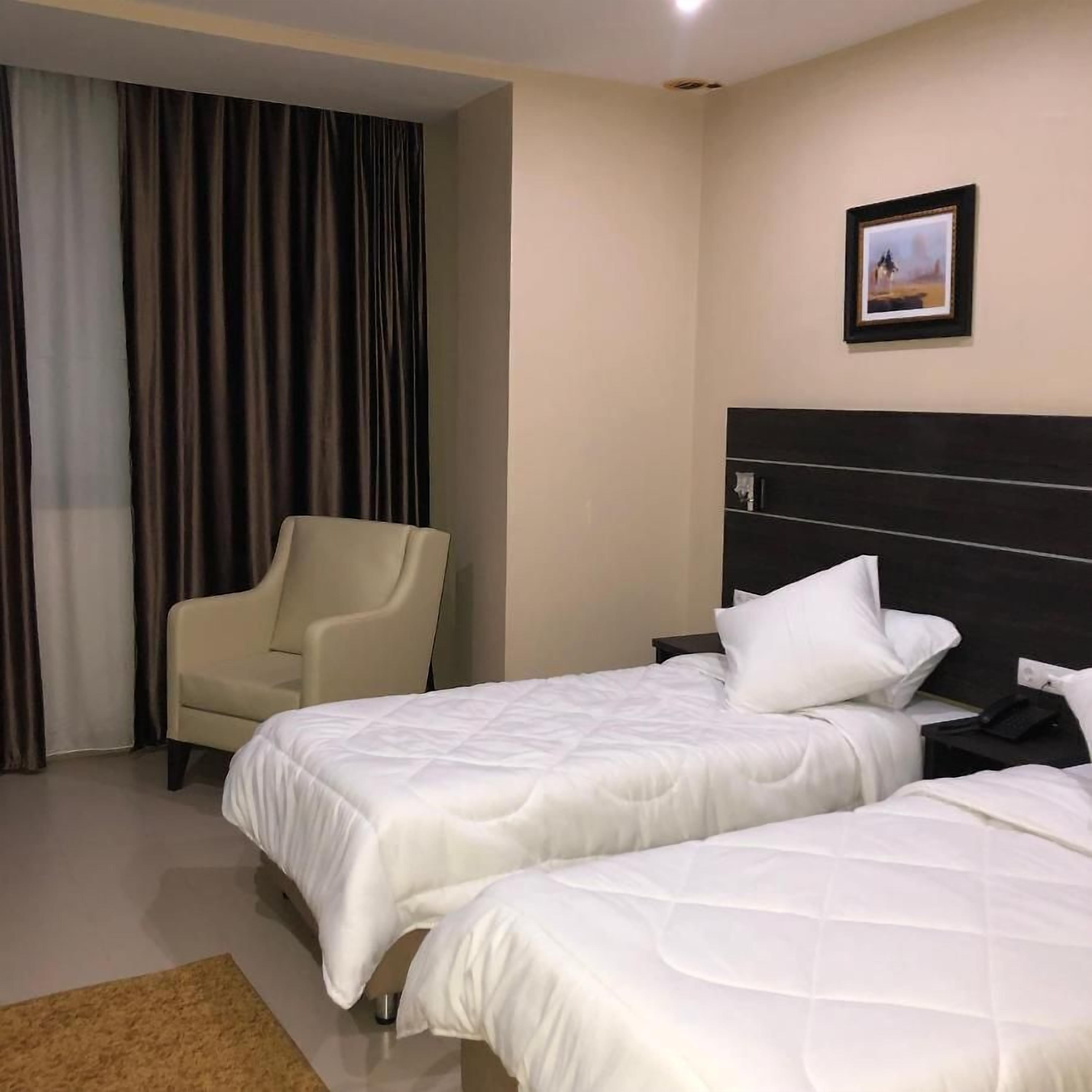Una habitación de hotel con dos camas, una silla, una mesita de noche y un cuadro en la pared.