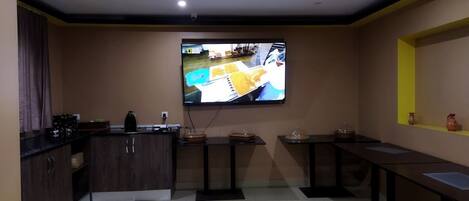 A modern kitchen with a flat-screen TV mounted on the wall, wooden cabinets, and a small dining area with tables and chairs.