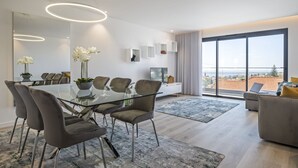 A modern living room with a glass dining table, grey upholstered chairs, a sofa, and a large window offering a view of the city.