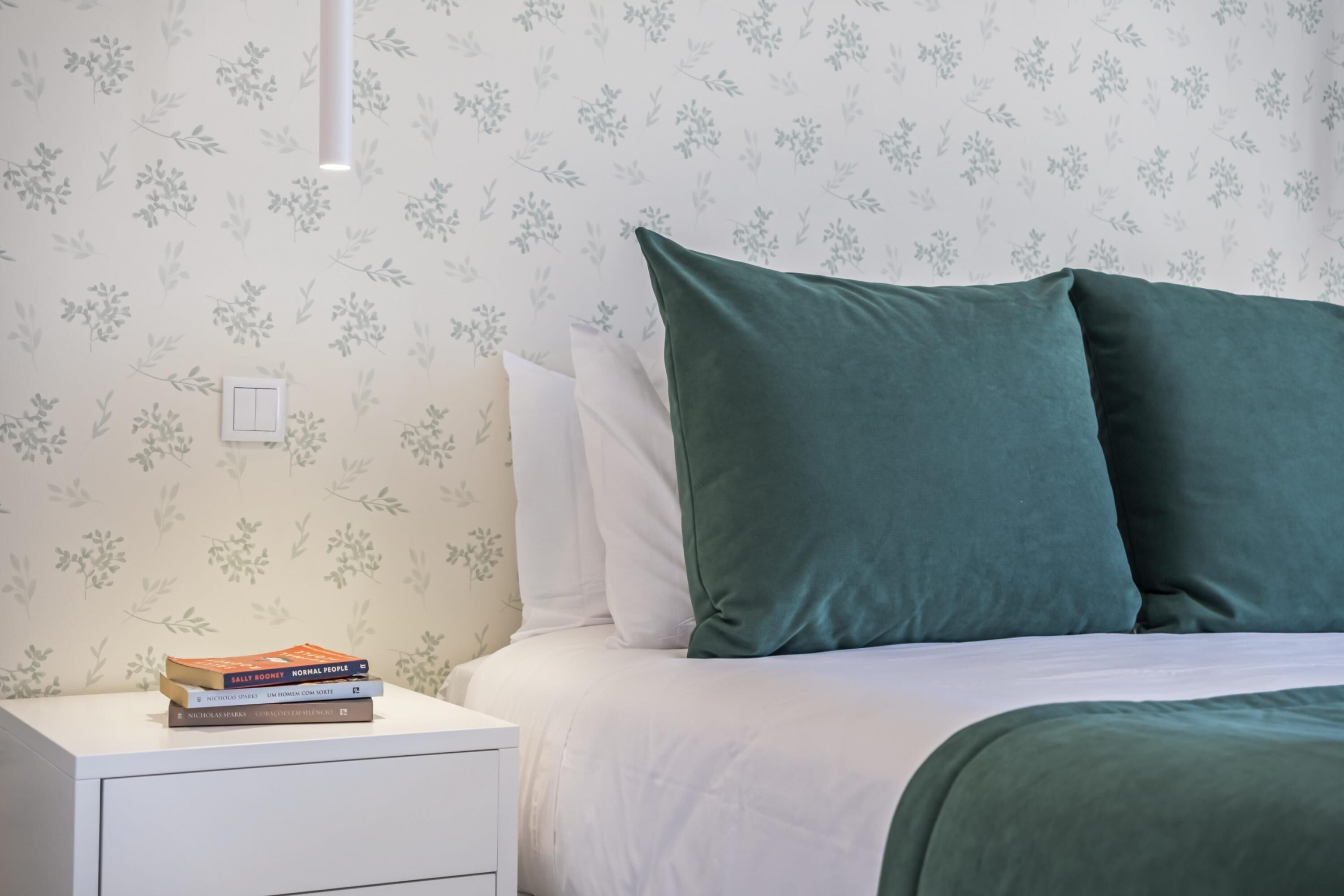 A neatly made bed with green pillows, a bedside table with a stack of books, and a wall with a floral pattern.