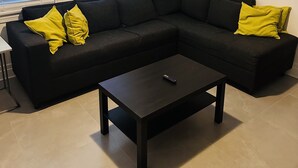 A living room with a black sectional sofa, two yellow pillows, a black coffee table, and a remote control on it.