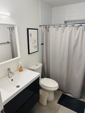 Un baño con un lavamanos blanco, un inodoro, una cita motivacional enmarcada, una cortina de ducha y una alfombra negra.