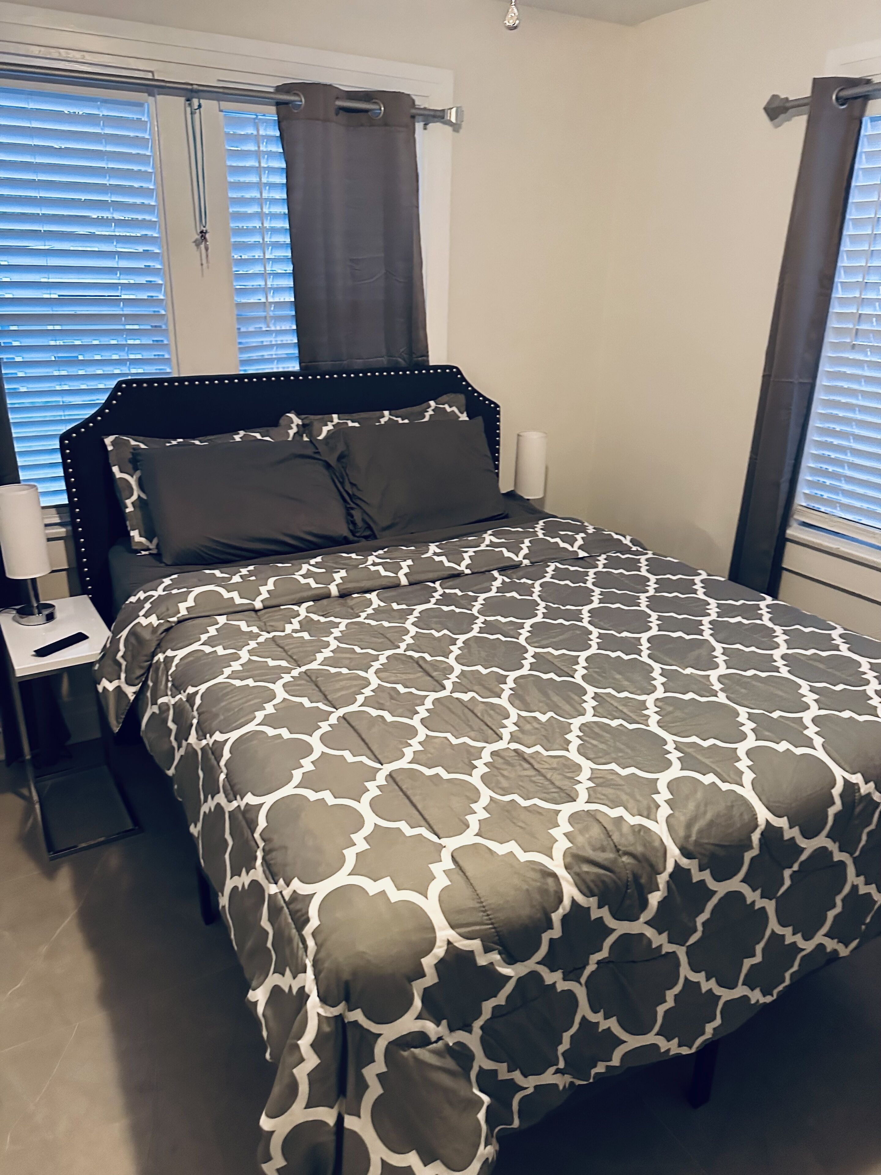 Ein ordentlich bezogenes Bett mit einem gemusterten Bettüberwurf, ein Nachttisch mit Lampe und ein Fenster mit Jalousien.