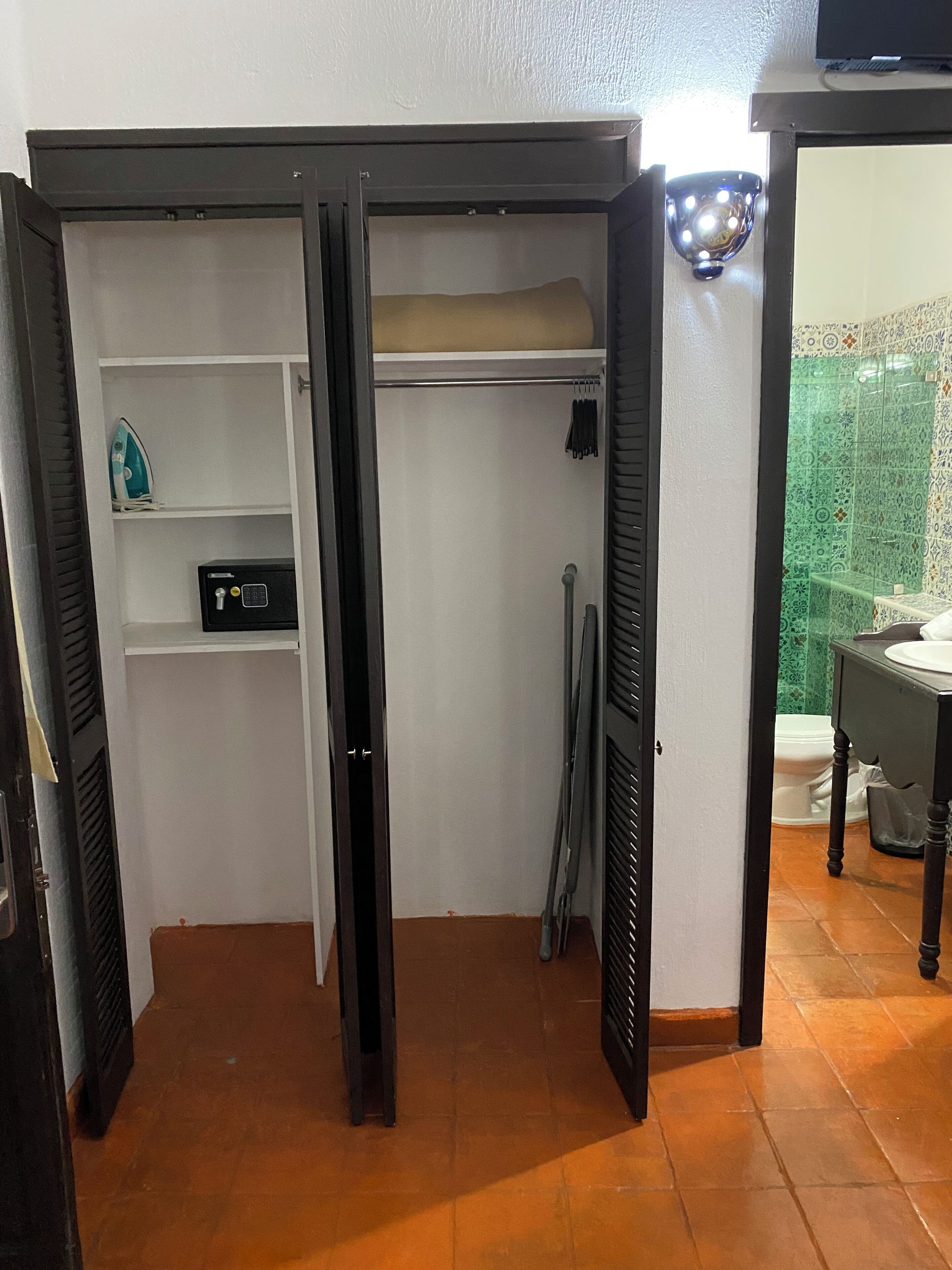 A room with a wardrobe, a shelf with an iron, a small black box, and a tiled floor.