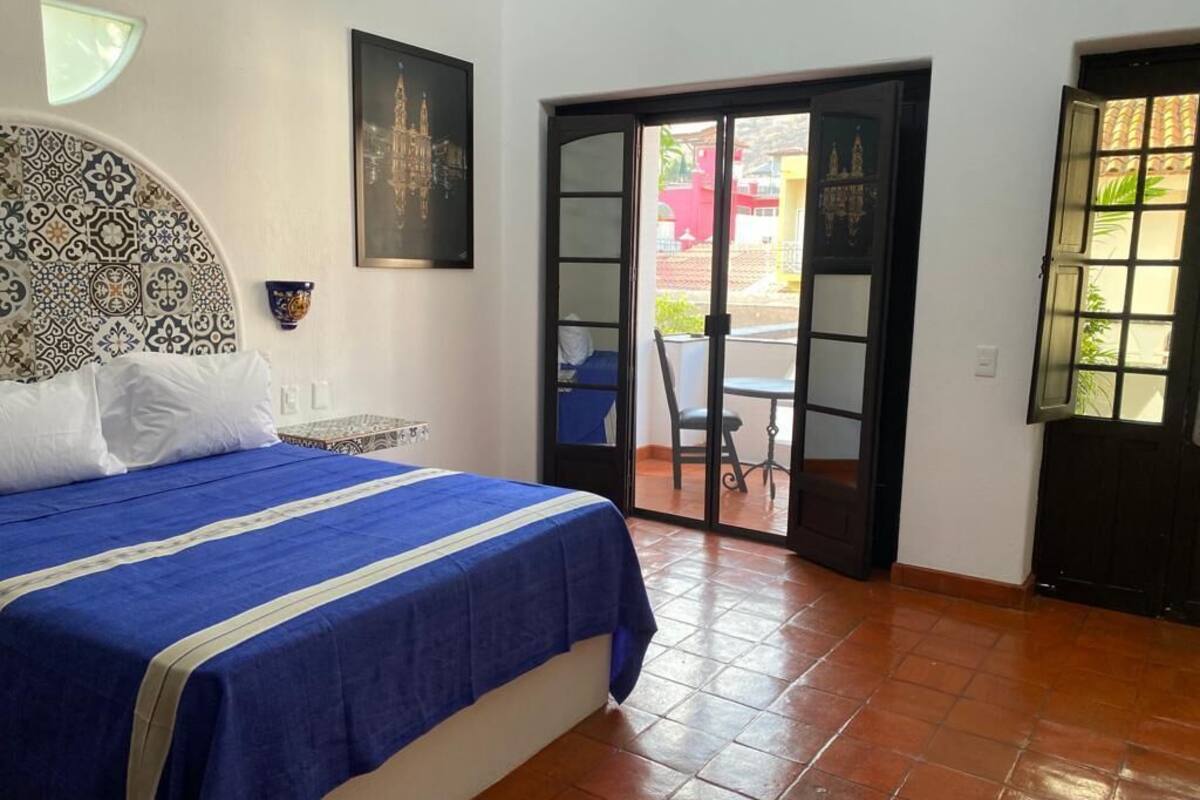 A bedroom with a patterned headboard, a bed with a blue bedspread, a wall-mounted clock, and a door leading to an outdoor area with a table and chairs.