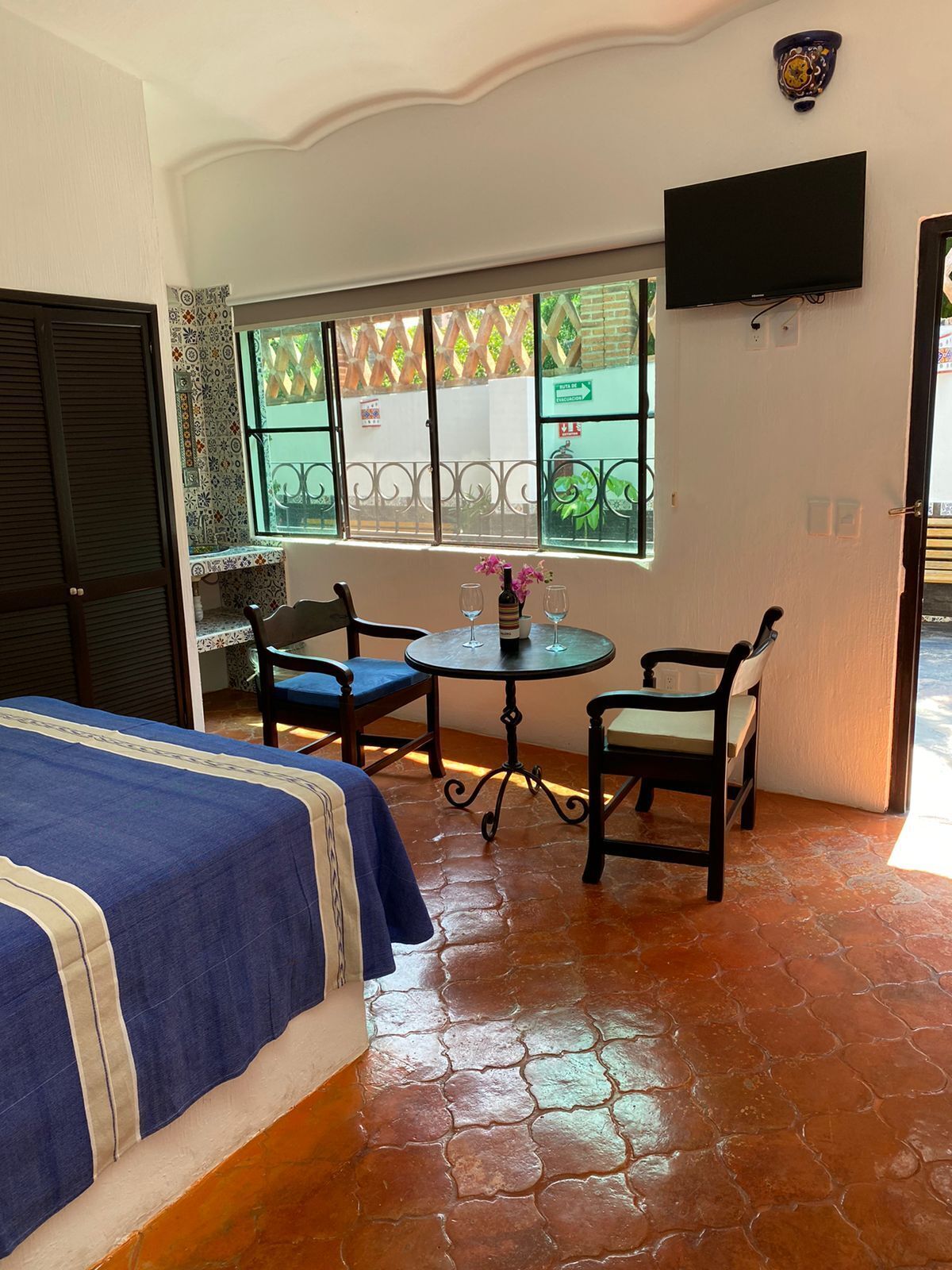 A room with a bed, two chairs, a small table with a bottle and glasses, a TV mounted on the wall, and a window with a view of greenery.