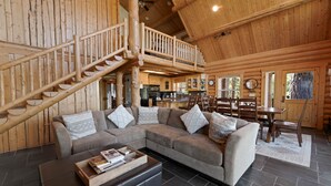 A log cabin interior with a wooden staircase, a large sectional sofa, a dining table, and a kitchen area.