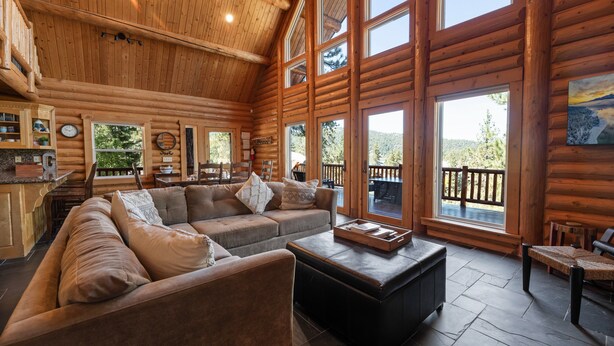 A log cabin interior with a large sofa, a coffee table, and a dining area with a view of the outdoors.